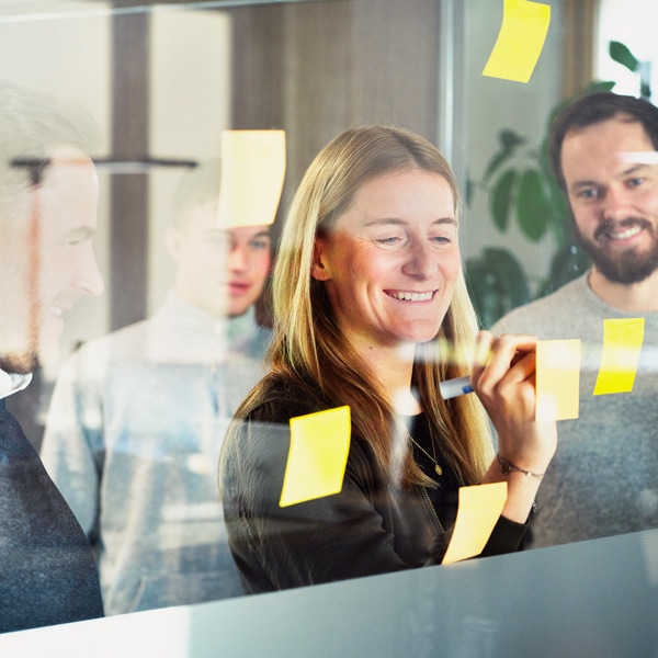 im Vordergrund stehen ein Mann und eine Frau aus dem Strategic Projects & Business Developtment Team an einer Glaswand. Sie führen dabei ein Gespräch und schreiben etwas auf Post-it Zetteln. 