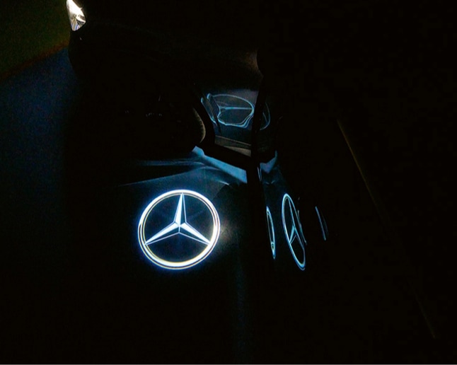  Ein beleuchtetes Mercedes-Benz Logo, das auf einen dunklen Untergrund projiziert wird. 
