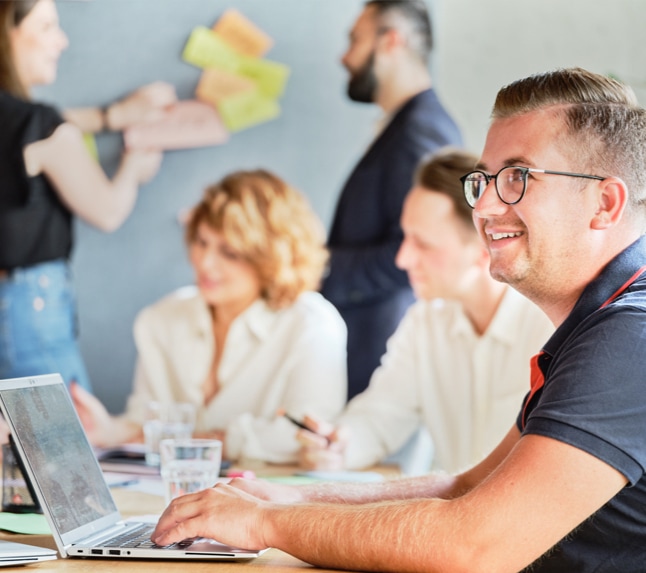 Mehrere Teammitglieder sitzen an einem Besprechungstisch. Die Person im Vordergrund sitzt an einem Laptop. Im Hintergrund sitzen zwei Personen, die sich offensichtlich unterhalten. 