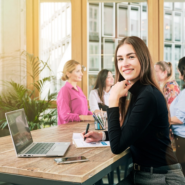 Eine Frau aus dem General Management Team sitzt vor einem Laptop, lächelt in die Kamera und hält einen Stift. Im Hintergrund unterhalten sich vier Frauen.