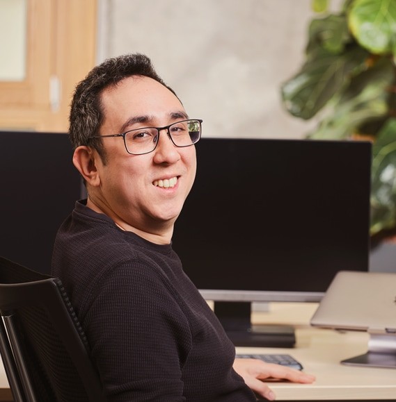 Ein Mann aus dem Informationstechnologie Team lächelt in die Kamera, während er an einem Schreibtisch mit zwei Monitoren und einem Laptop sitzt.